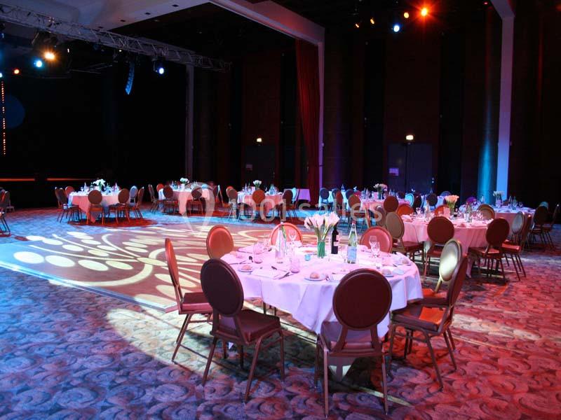 Salle de réception avec tables rondes dressées, éclairage coloré et ambiance festive.
