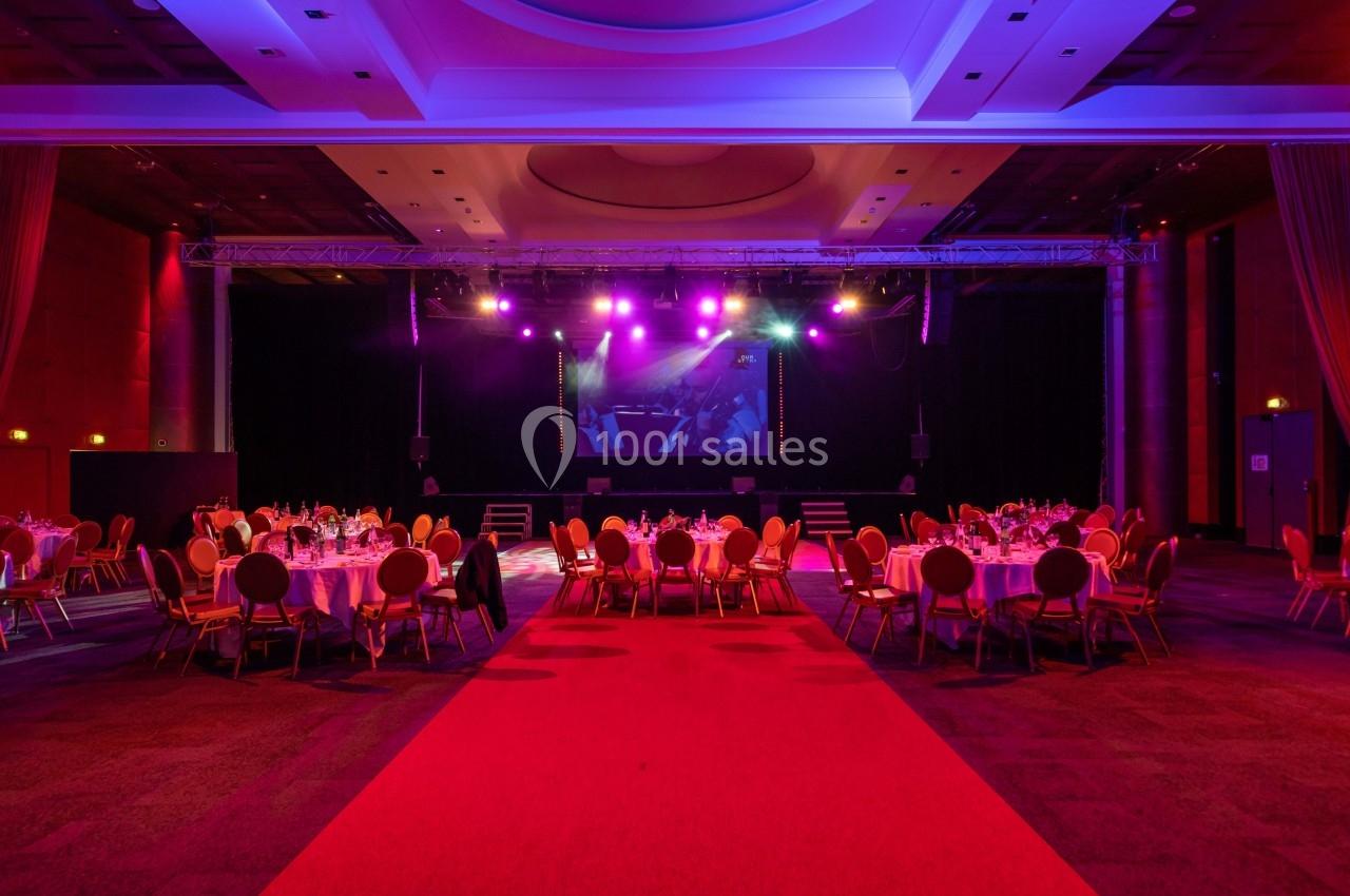 Salle de réception avec tables rondes dressées, éclairage coloré et scène en fond sous des projecteurs.