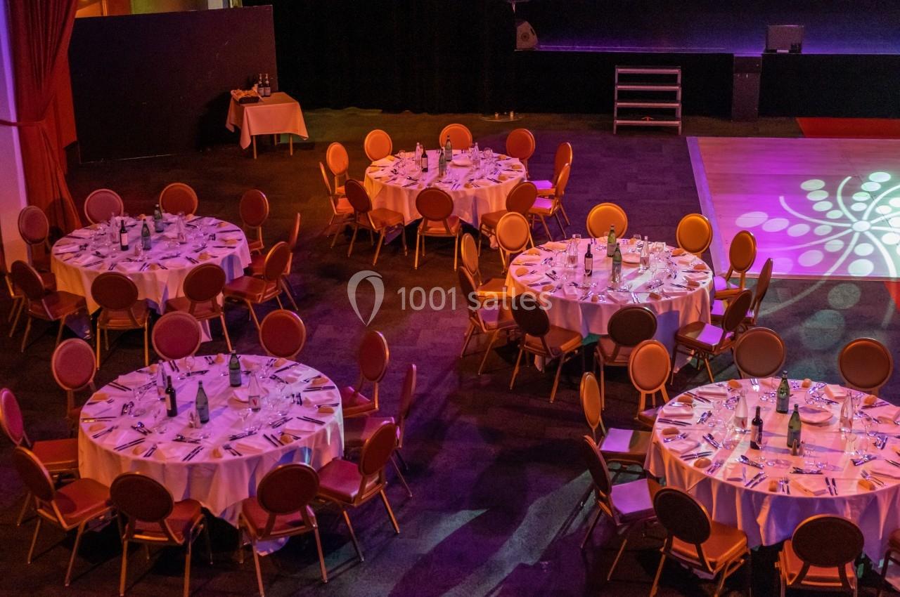 Salle de réception avec tables rondes dressées, nappes blanches, chaises disposées et éclairage coloré.