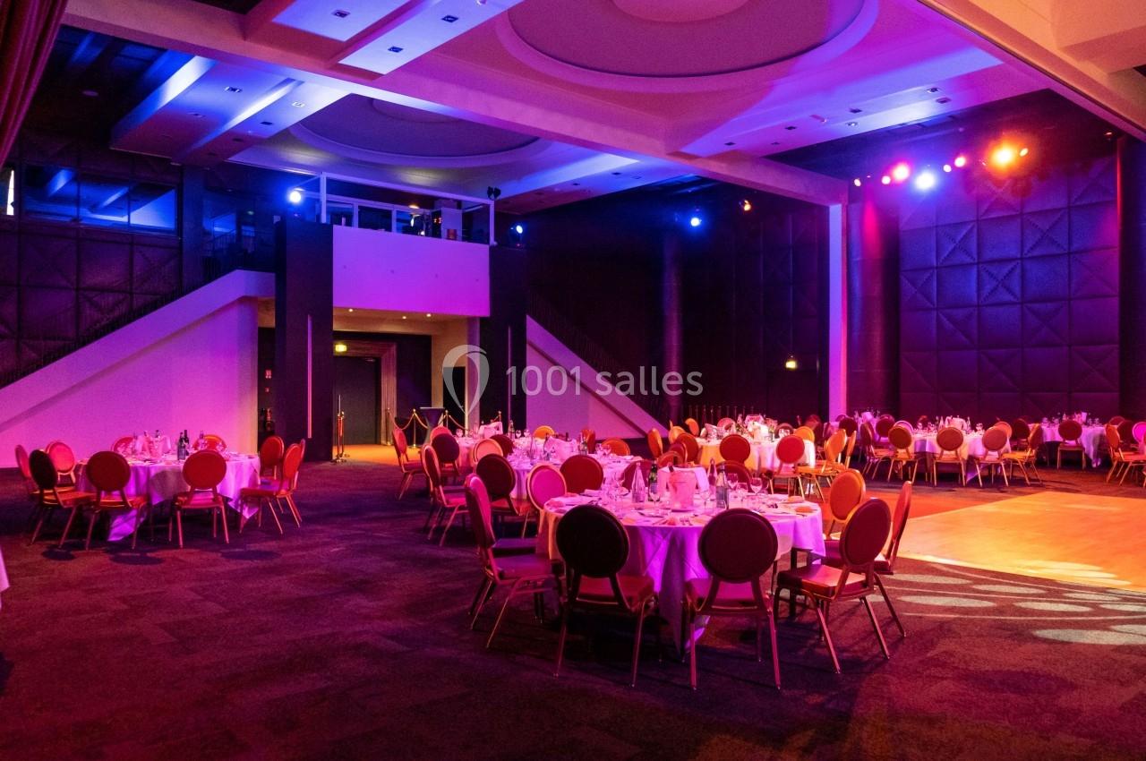 Salle de réception avec tables rondes dressées, éclairage coloré et scène illuminée en arrière-plan.