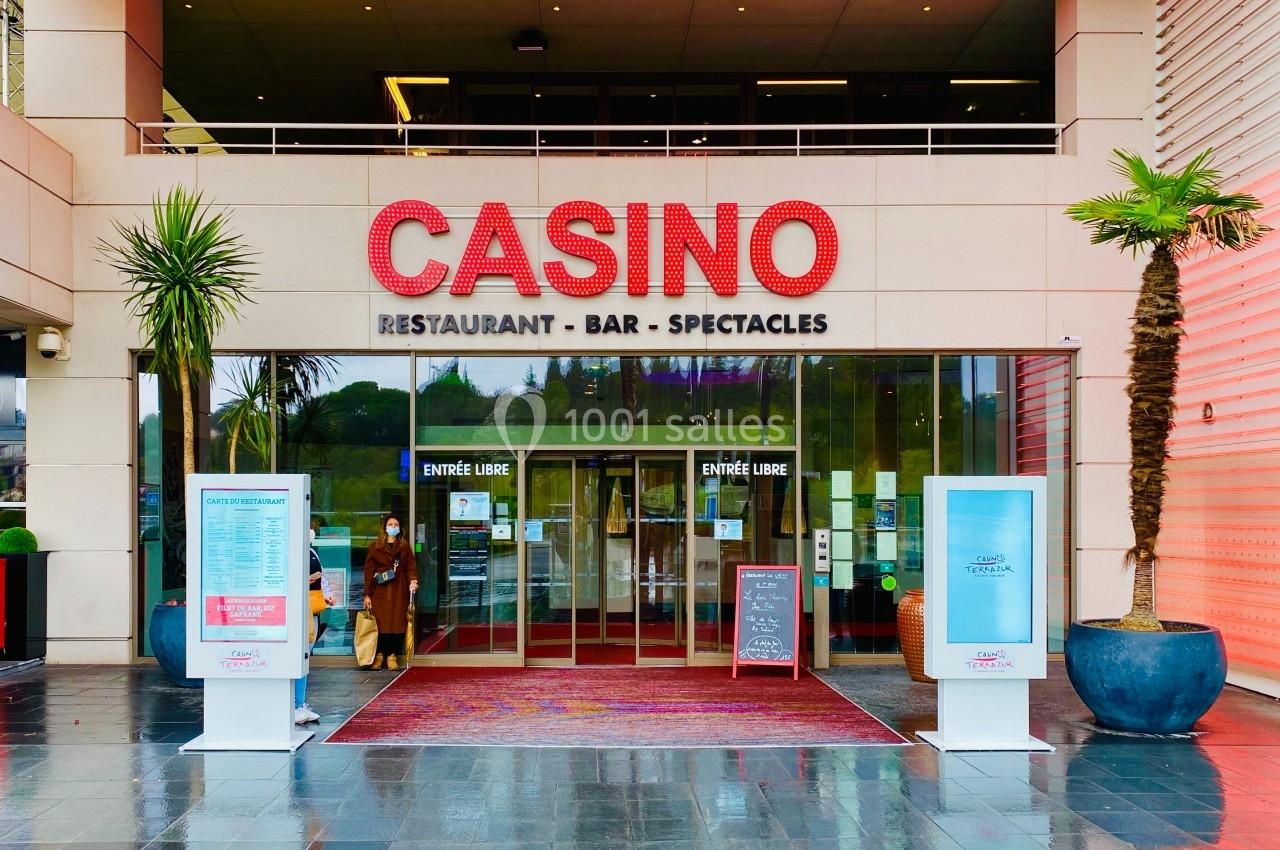 Entrée principale d'un casino avec enseigne lumineuse rouge, affiches d'information et palmiers décoratifs.