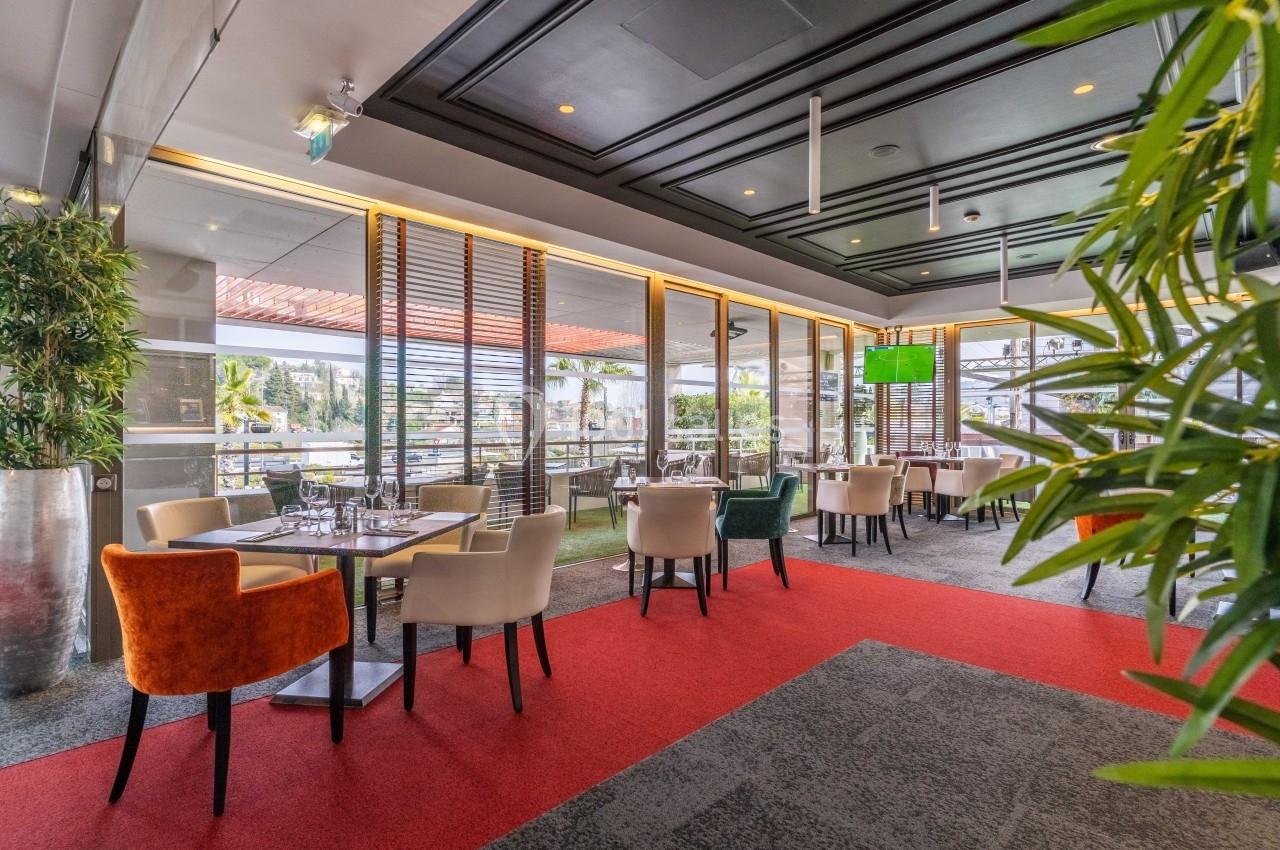 Salle de restaurant lumineuse avec grandes baies vitrées, tables dressées et chaises colorées, vue sur l'extérieur.