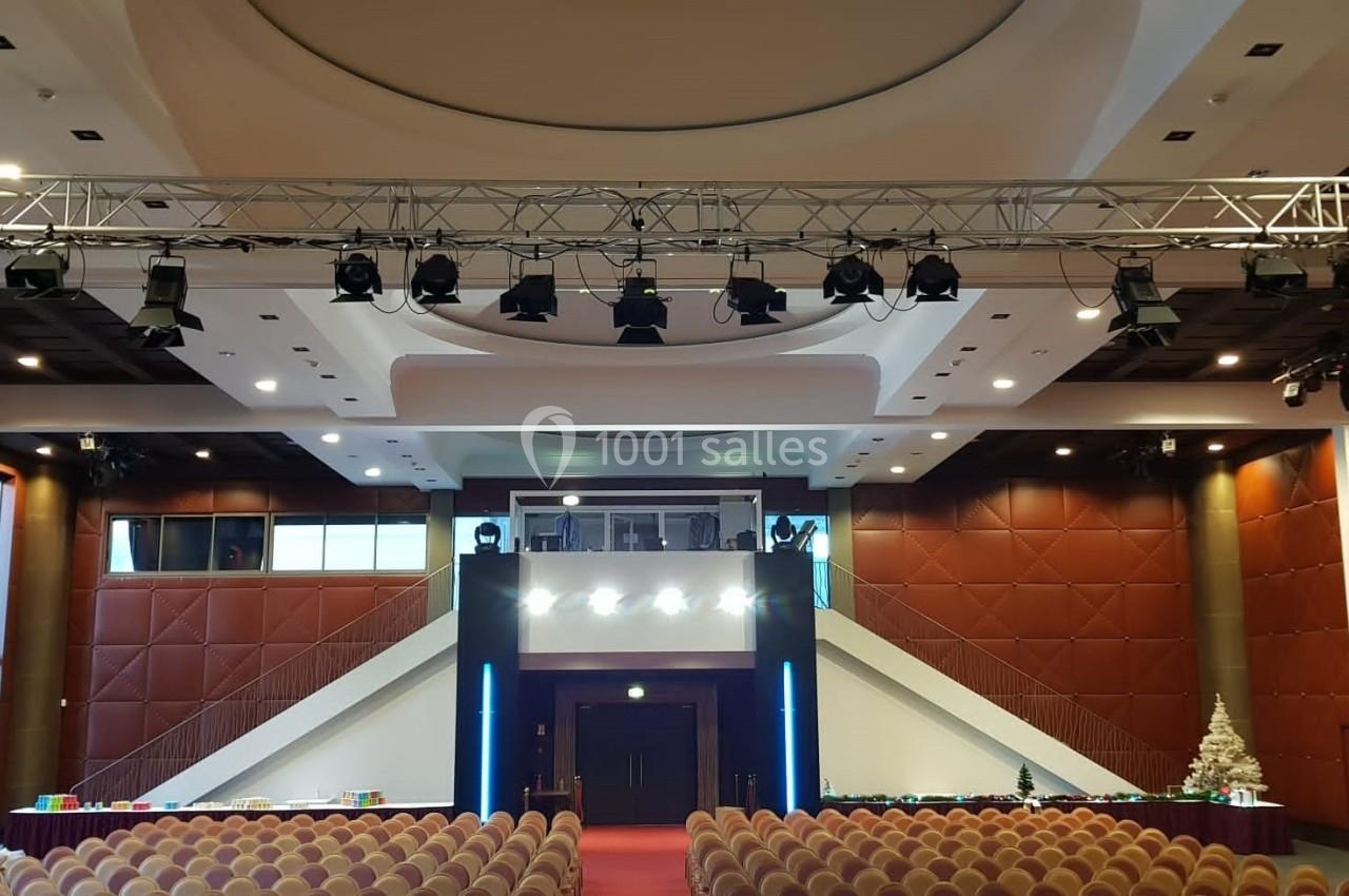 Salle de conférence avec sièges alignés, scène centrale, éclairages suspendus et sapin décoratif à droite.