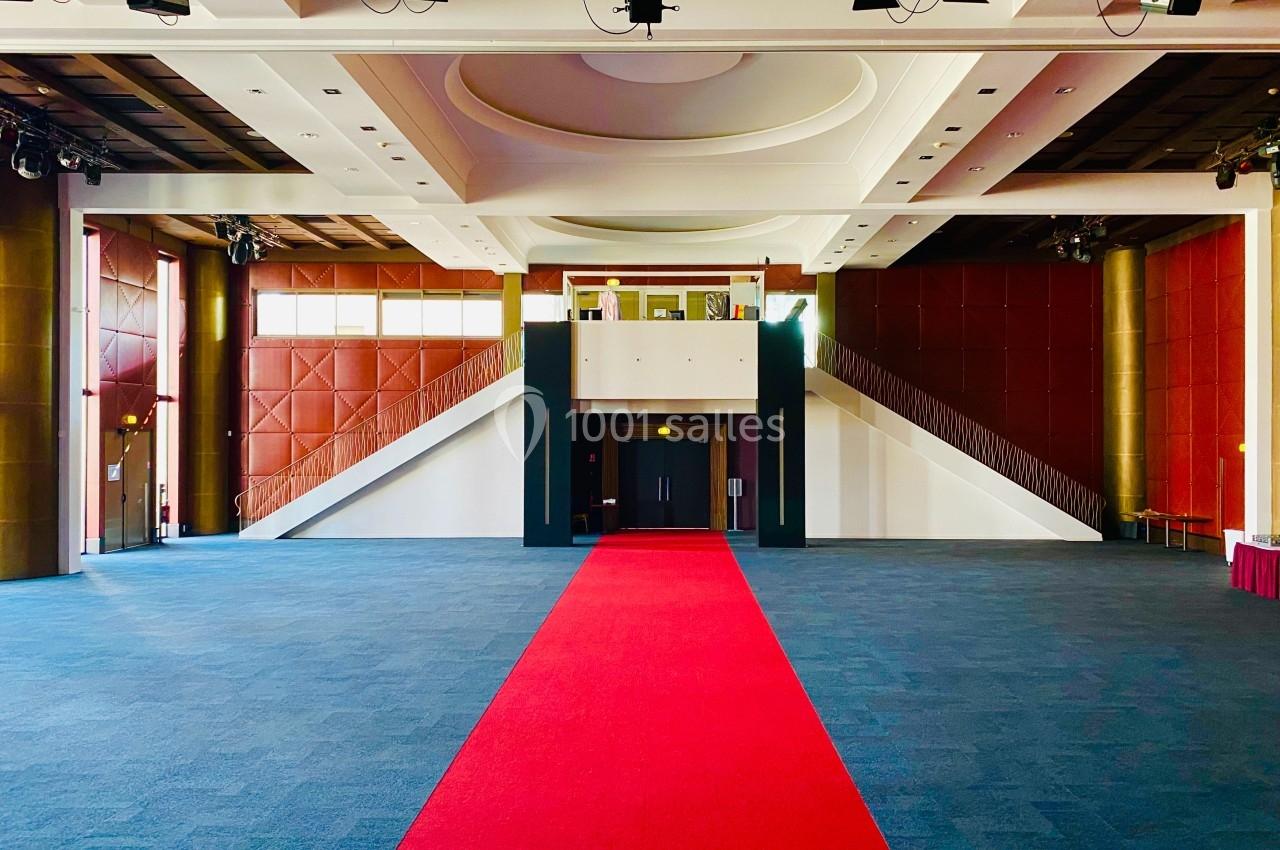 Grande salle vide avec moquette bleue, tapis rouge central menant à un escalier symétrique et plafond lumineux.