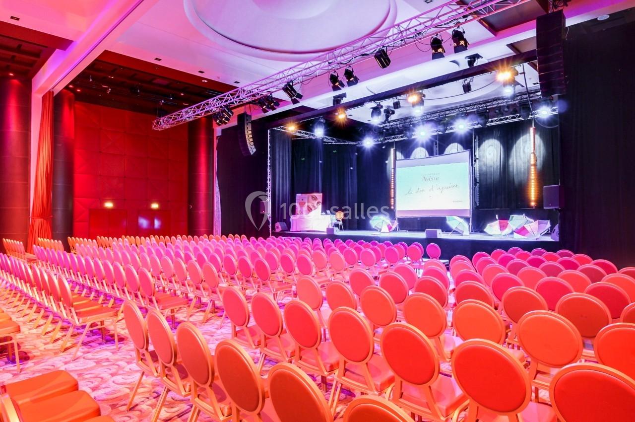 Salle de conférence avec des rangées de chaises vides, éclairage coloré et écran de projection sur scène.