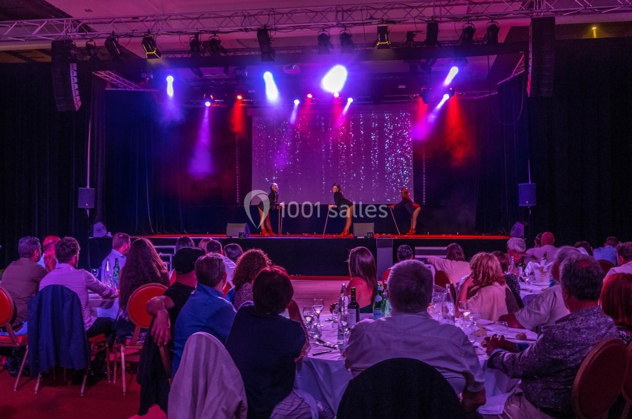 Spectacle sur scène avec trois danseurs sous des lumières colorées, devant un public assis à des tables.