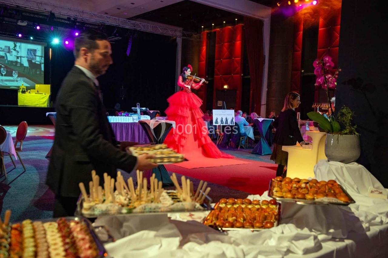 Buffet de plats variés au premier plan, avec une violoniste en robe rouge jouant sur une scène éclairée en arrière-plan.