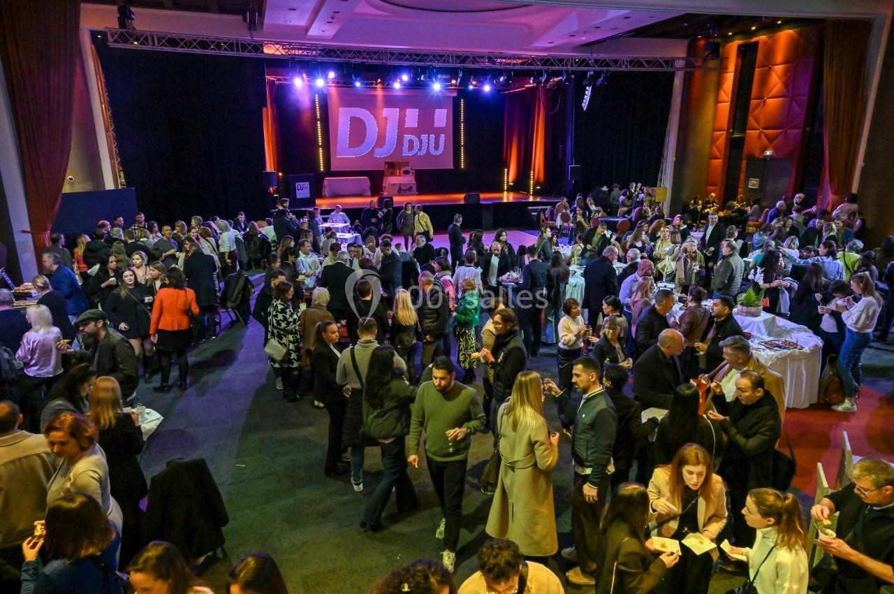 Salle comble avec des personnes debout et discutant lors d'un événement, une scène éclairée avec un DJ en arrière-plan.