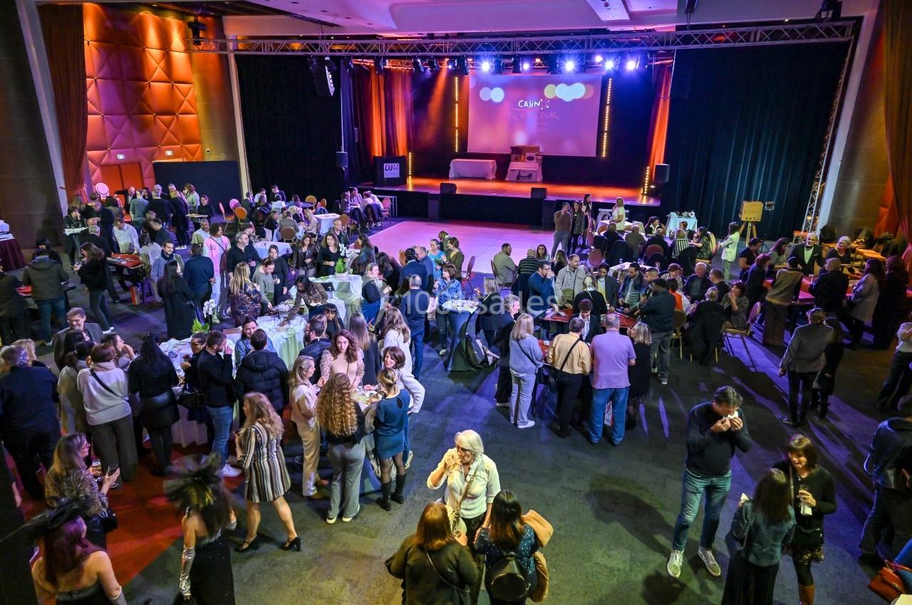 Salle de réception animée avec des groupes de personnes discutant et un espace scénique éclairé en arrière-plan.