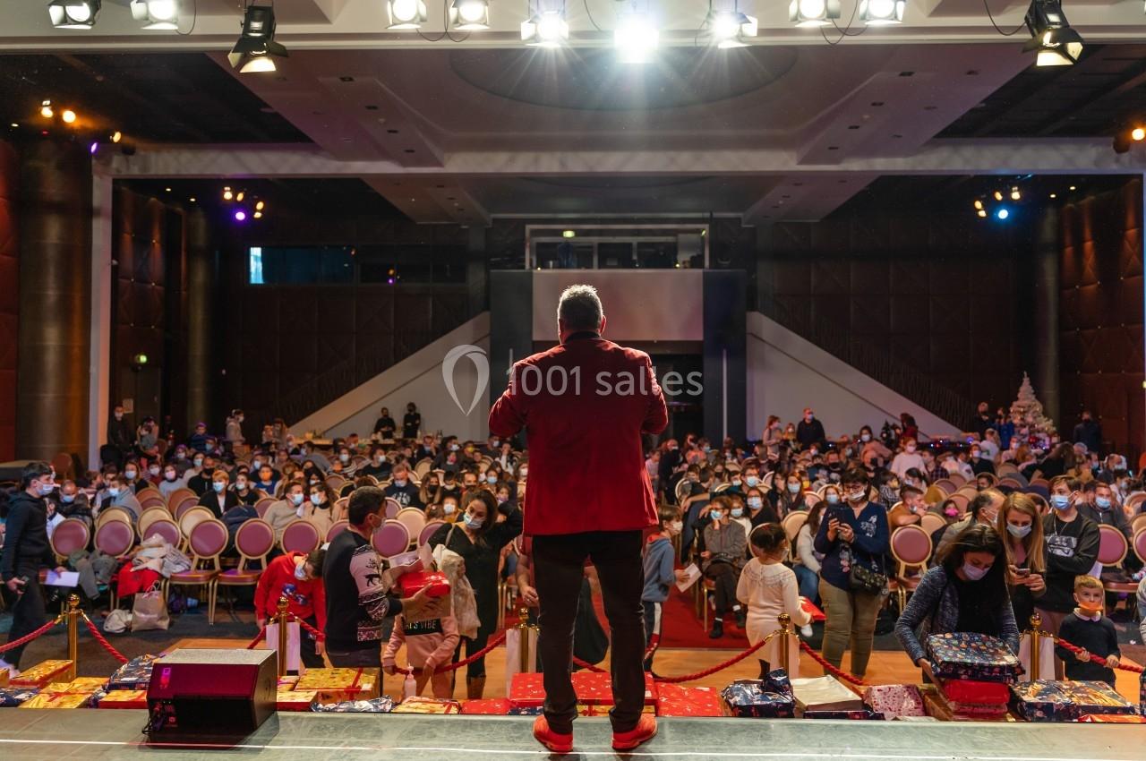 Un homme en veste rouge s'adresse à un public assis dans une grande salle éclairée, avec des enfants au premier plan.