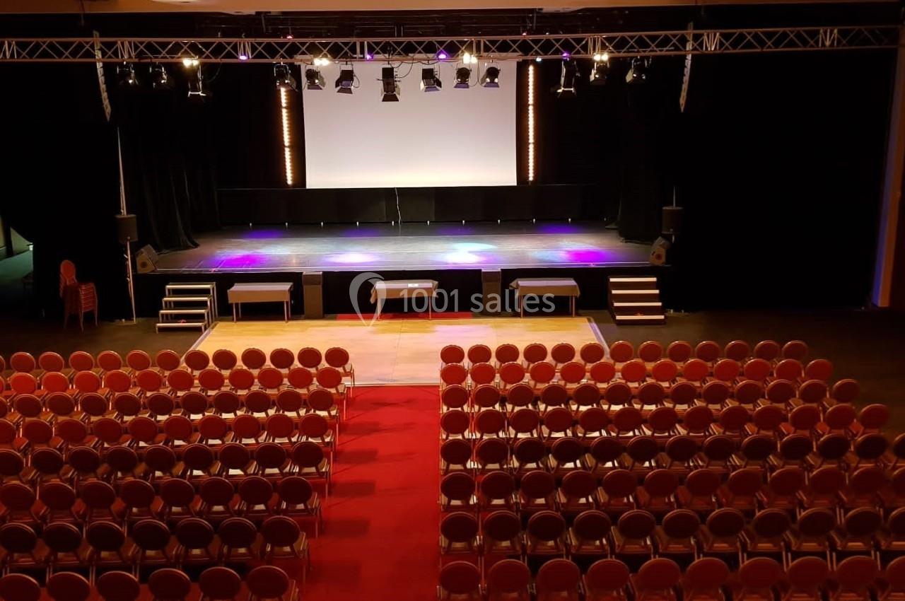 Salle de spectacle avec scène éclairée, rangées de sièges rouges et allée centrale menant à la scène.