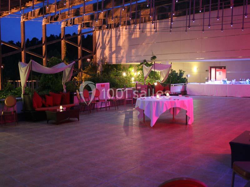 Terrasse éclairée en soirée avec des tables dressées, des canapés et une décoration lumineuse sous une structure en bois.