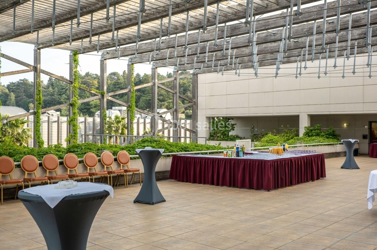 Terrasse aménagée avec des tables hautes, une longue table de buffet et des chaises alignées, entourée de verdure.