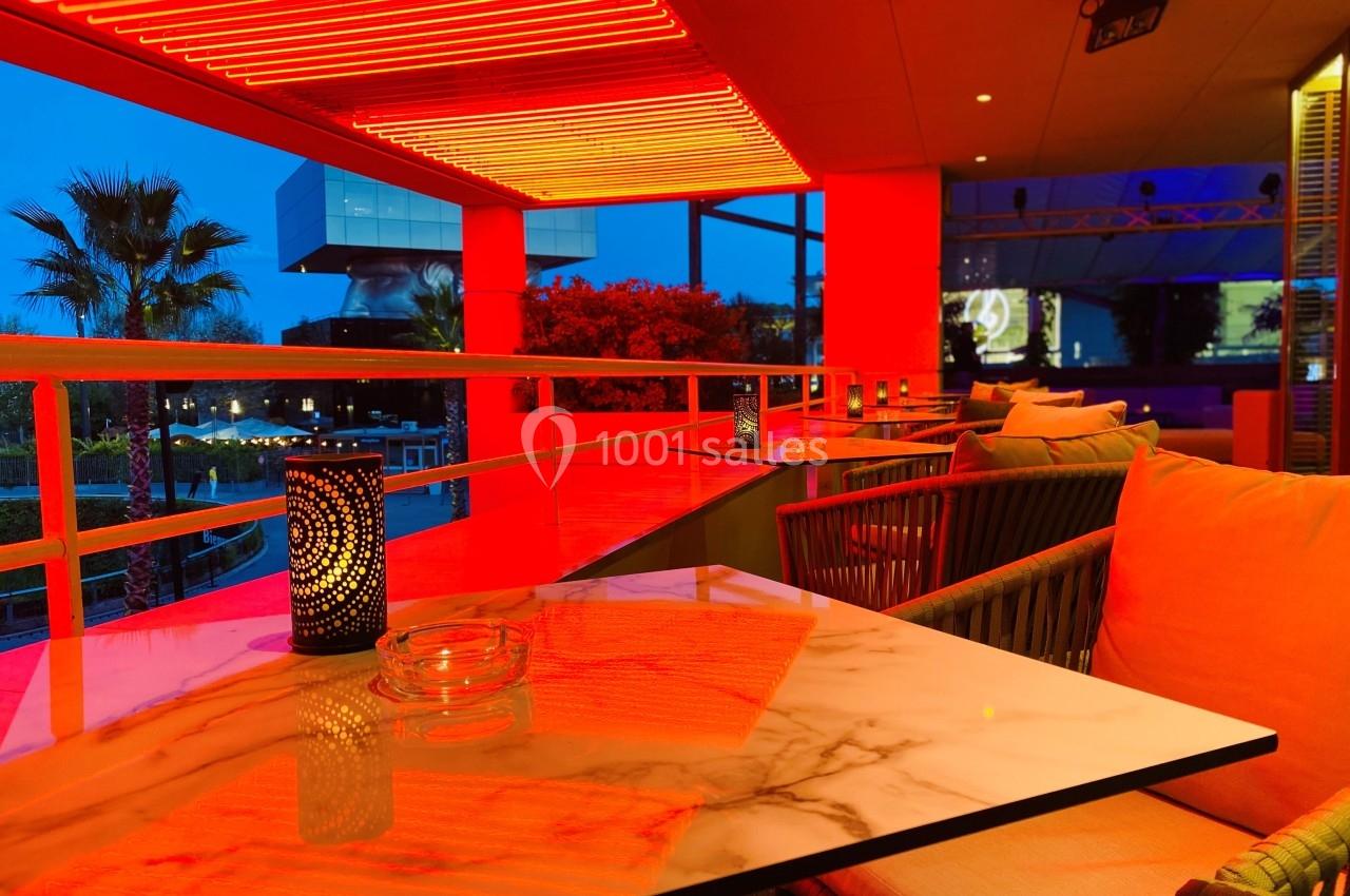 Terrasse moderne éclairée par des lumières rouges, avec tables en marbre, chaises confortables et vue sur un extérieur…
