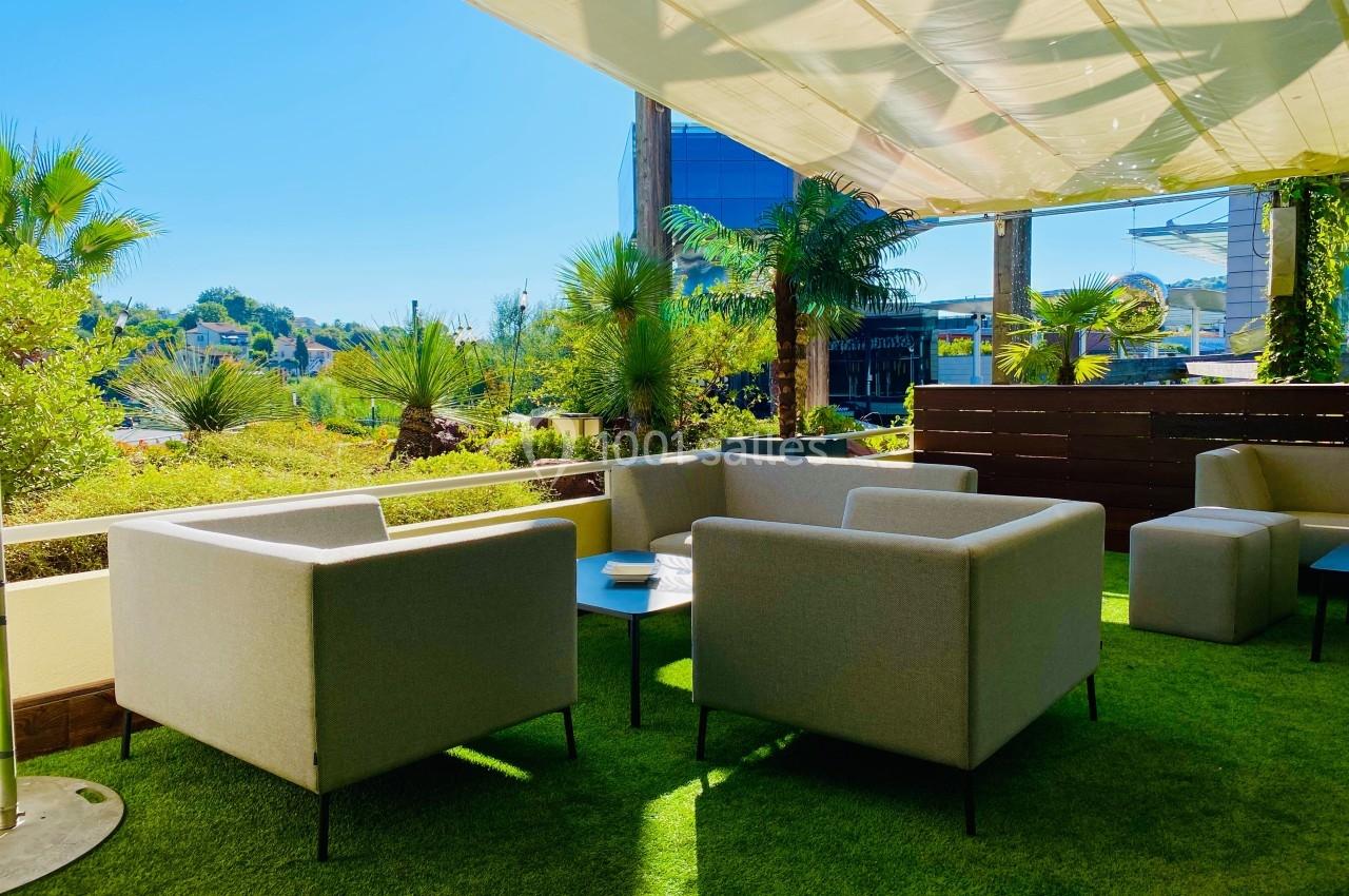 Espace extérieur avec fauteuils gris, tables basses et pelouse artificielle, ombragé par une toile tendue.