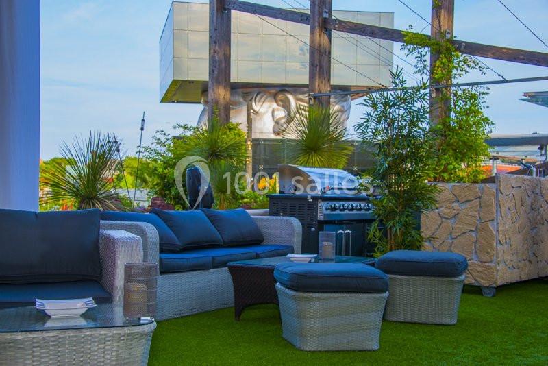 Salon de jardin avec canapés en rotin, barbecue, plantes et pelouse artificielle sur une terrasse extérieure.