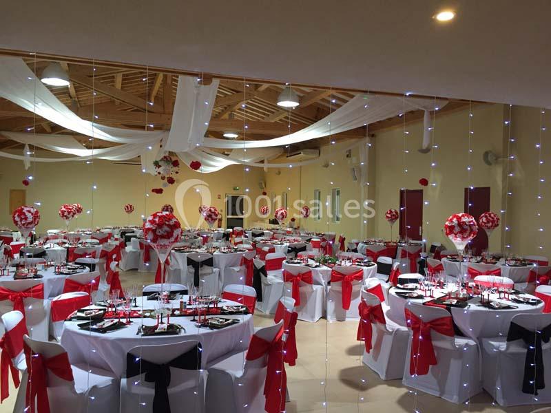 Salle de réception décorée avec des tables rondes, nappes blanches, nœuds rouges et noirs, et guirlandes lumineuses.