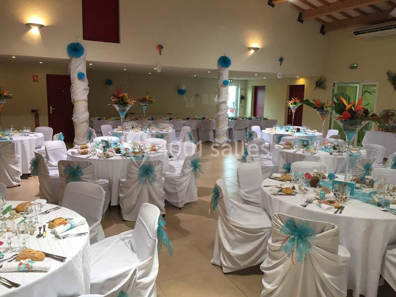 Salle de réception décorée avec des tables rondes, nappes blanches, chaises ornées de rubans bleus et arrangements floraux.