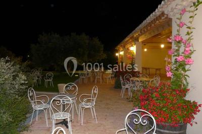 Terrasse extérieure éclairée la nuit, avec des tables et chaises en fer forgé entourées de fleurs colorées.