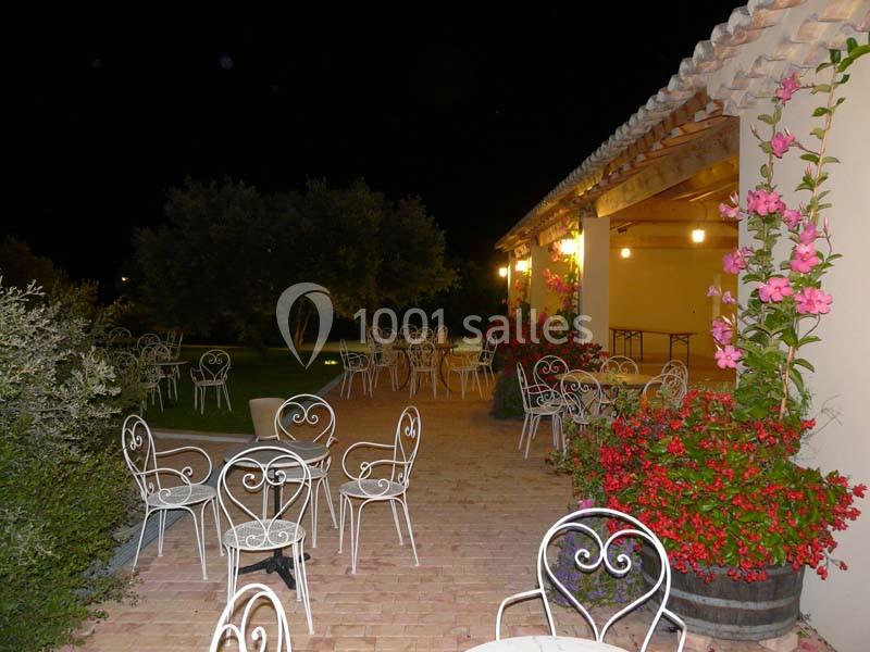 Terrasse extérieure éclairée de nuit avec des tables et chaises en fer forgé, entourée de fleurs et de verdure.