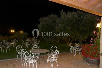 Terrasse extérieure éclairée la nuit, avec des tables et chaises en fer forgé entourées de fleurs colorées.