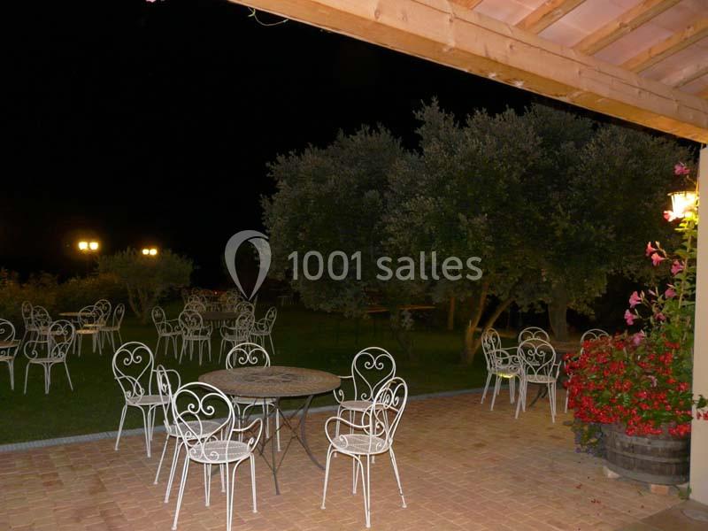 Terrasse extérieure avec tables et chaises en fer forgé, éclairée par des lampes, entourée d'arbres et de fleurs.