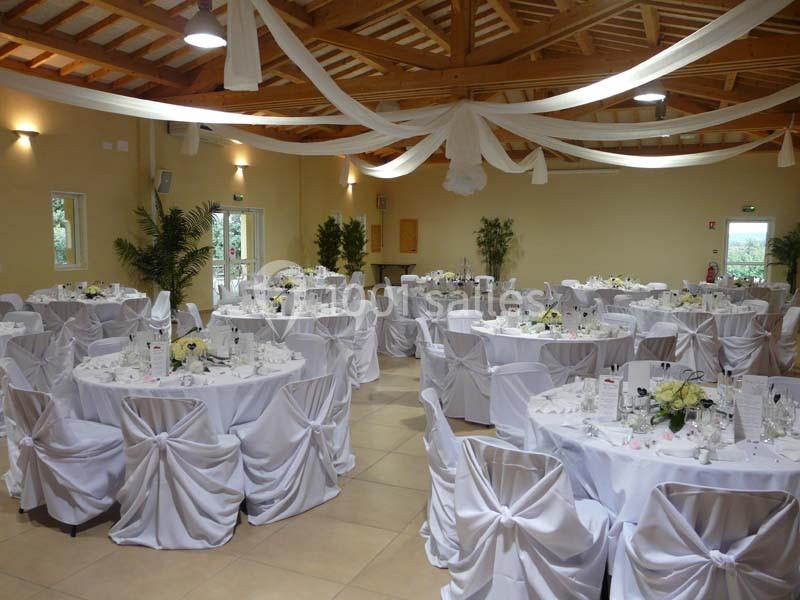 Salle de réception décorée avec des tables rondes, nappes blanches et chaises couvertes, sous un plafond en bois.
