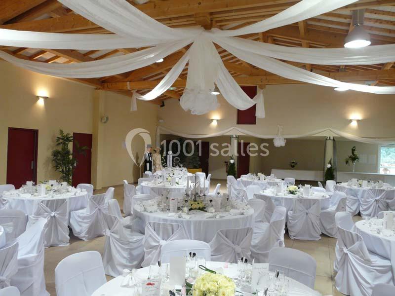Salle de réception décorée pour un mariage, avec des tables rondes dressées et des drapés blancs suspendus au plafond.