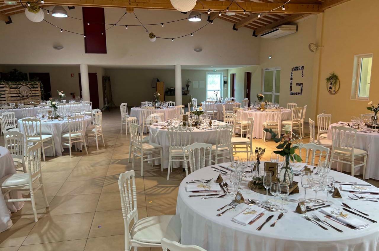 Salle de réception décorée avec des tables rondes dressées, chaises blanches et guirlandes lumineuses suspendues.