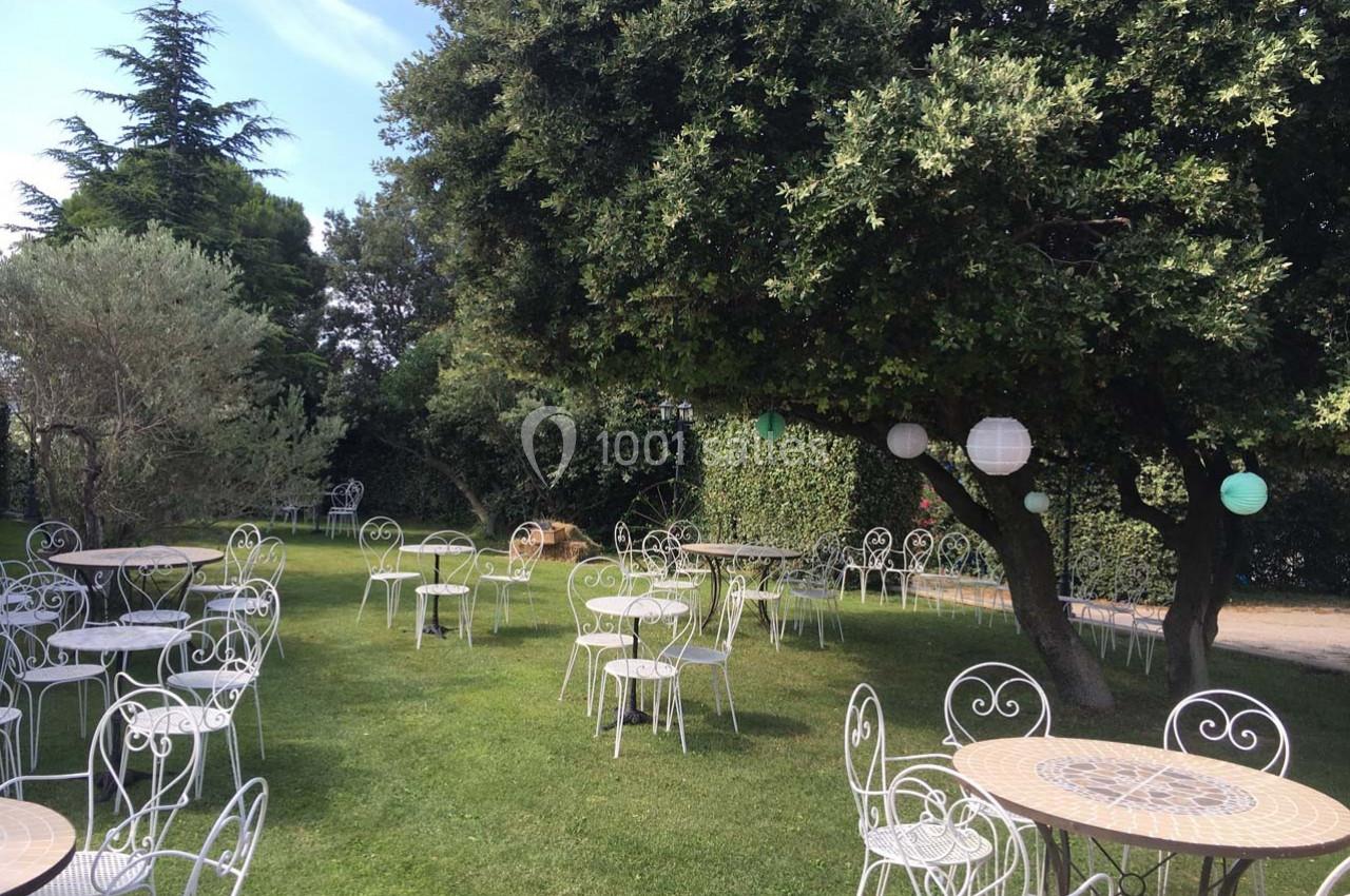 Chaises et tables en fer forgé disposées sur une pelouse, entourées d'arbres et de lanternes suspendues.