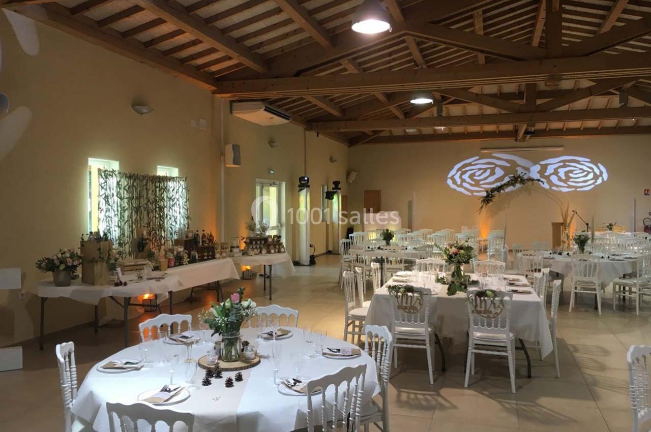 Salle de réception décorée pour un événement, avec tables dressées, chaises blanches et éclairage chaleureux.