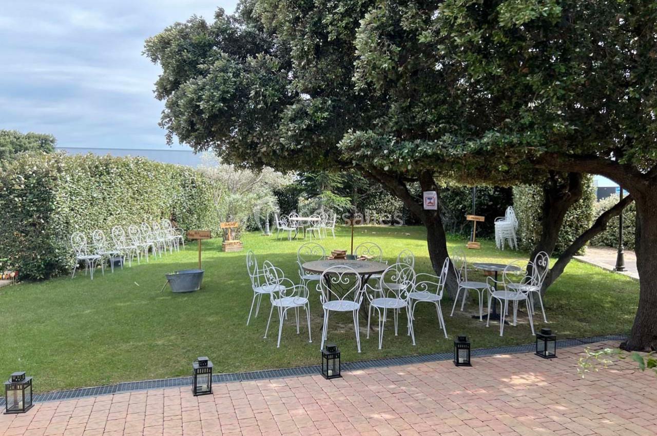 Jardin aménagé avec des tables et chaises blanches en fer forgé, sous des arbres et entouré de haies.
