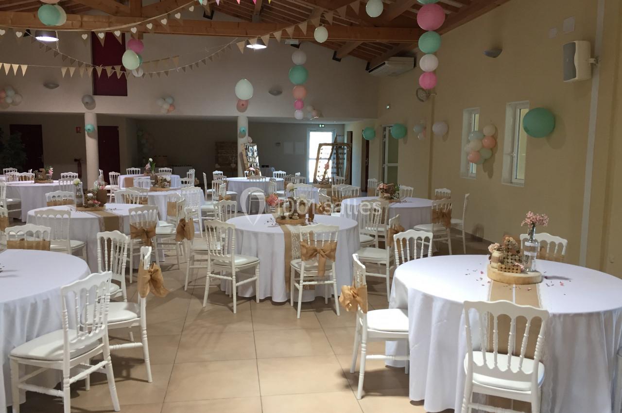 Salle décorée pour un événement avec des tables rondes nappées de blanc, chaises blanches et guirlandes colorées suspendues.