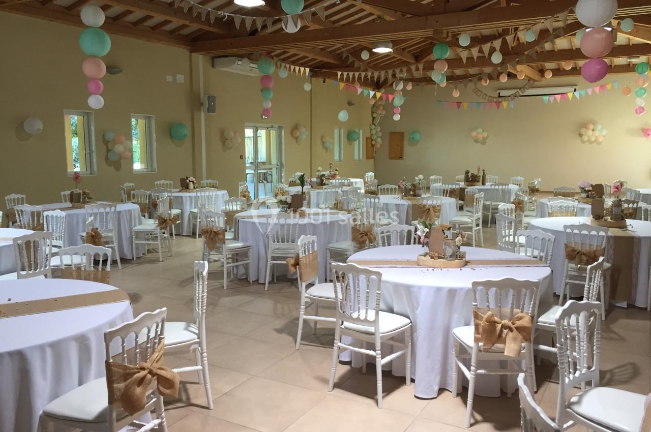 Salle décorée pour un événement, avec des tables rondes, nappes blanches, chaises décorées et guirlandes suspendues.