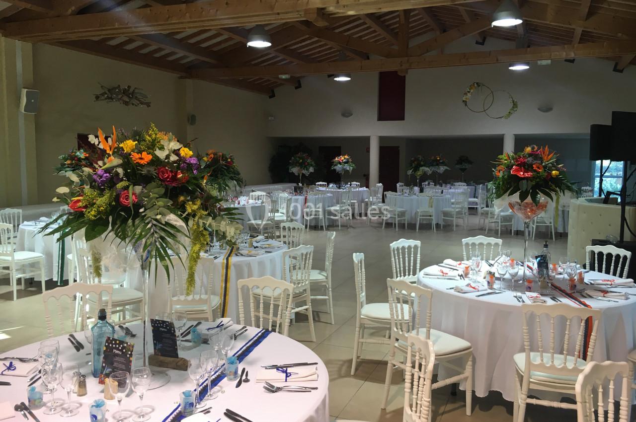 Salle de réception décorée avec des tables rondes dressées, ornées de bouquets de fleurs colorées.
