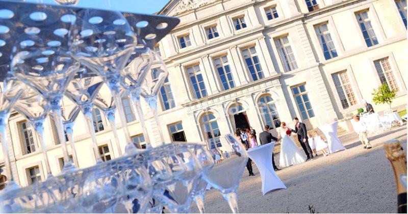 Des invités rassemblés devant un bâtiment historique lors d'une réception en extérieur, avec des verres empilés au premier…