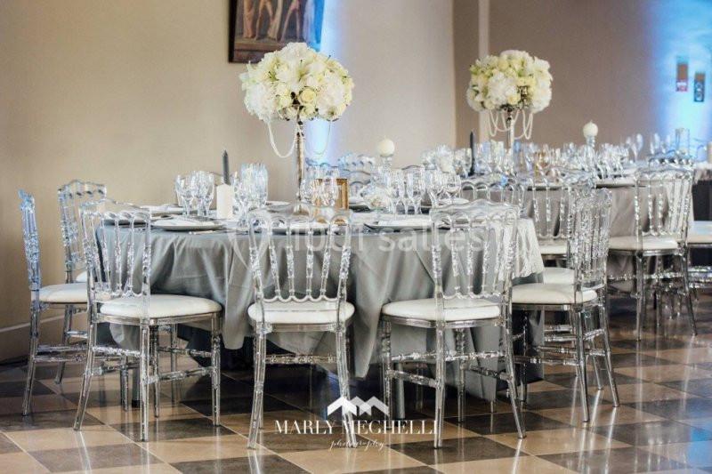 Tables élégamment dressées avec nappes grises, chaises transparentes et centres de table ornés de fleurs blanches.