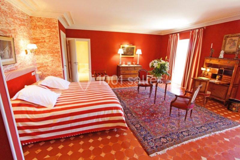 Chambre lumineuse avec murs rouges, lit à rayures, tapis oriental, meubles en bois et grande fenêtre avec rideaux.
