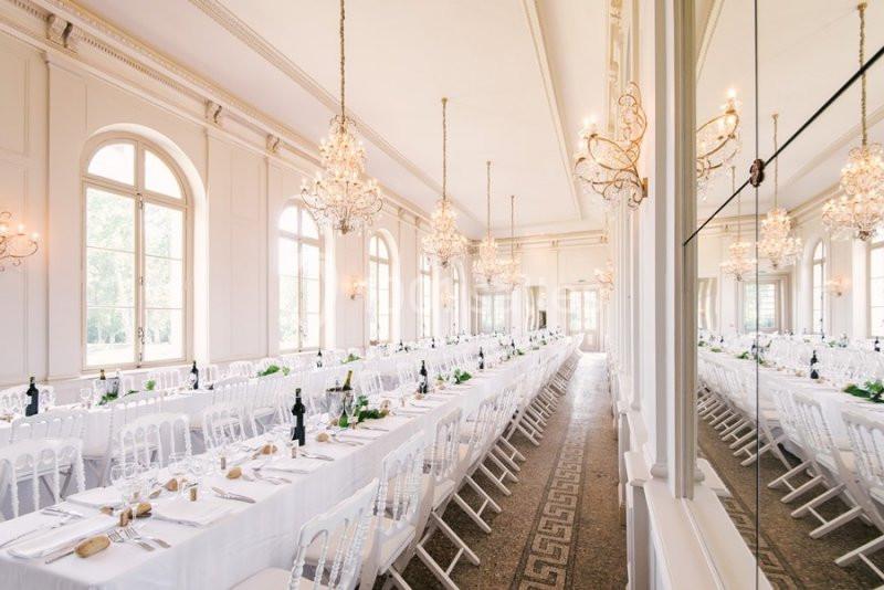 Salle de réception lumineuse avec de longues tables dressées, décorée de lustres et de moulures élégantes.