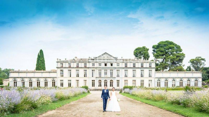 Un couple de mariés marche sur une allée bordée de fleurs devant un grand bâtiment historique sous un ciel bleu.