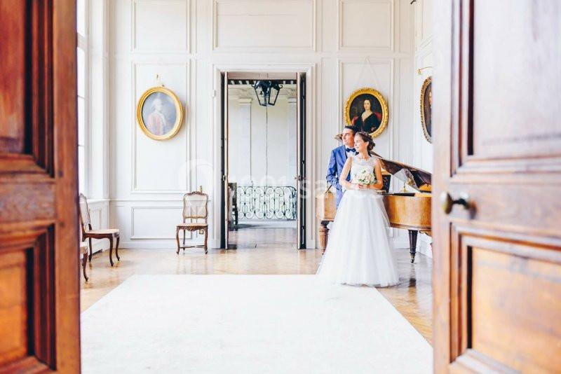 Une mariée en robe blanche et un homme posent près d'un piano dans une pièce lumineuse au décor classique.