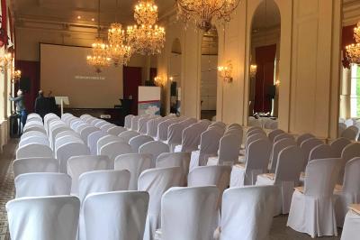 Salle de conférence élégante avec rangées de chaises blanches, lustres suspendus et écran de projection à l'arrière.