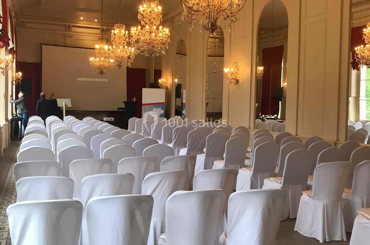 Salle de conférence élégante avec rangées de chaises blanches, lustres suspendus et écran de projection à l'arrière.