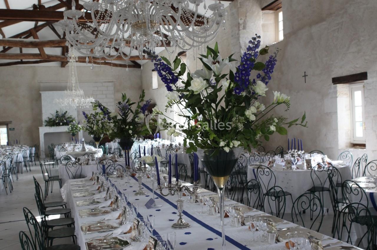 Salle de réception décorée avec une longue table dressée, ornée de fleurs blanches et violettes sous des lustres en cristal.