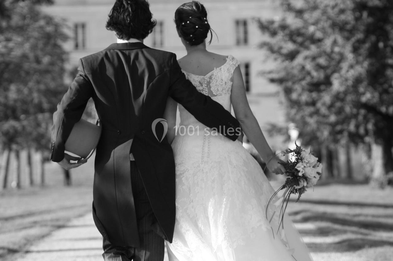Un couple de mariés marchant sur une allée bordée d'arbres, vu de dos, devant un bâtiment élégant.