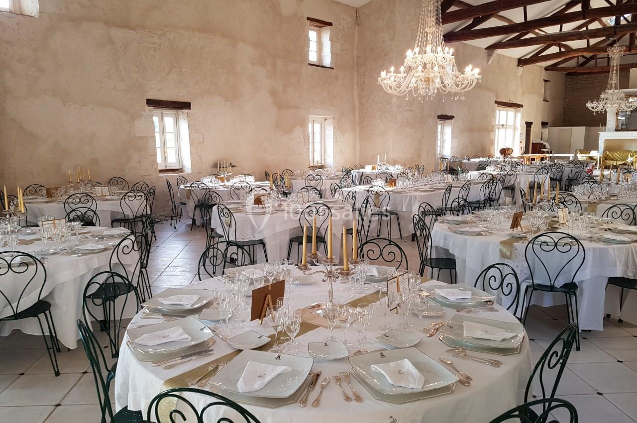 Salle de réception décorée avec des tables rondes dressées, nappes blanches, chandeliers dorés et chaises en fer forgé.