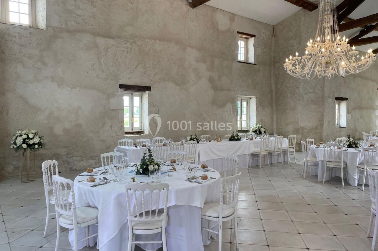 Salle de réception lumineuse avec tables rondes et rectangulaires décorées de nappes blanches et arrangements floraux.