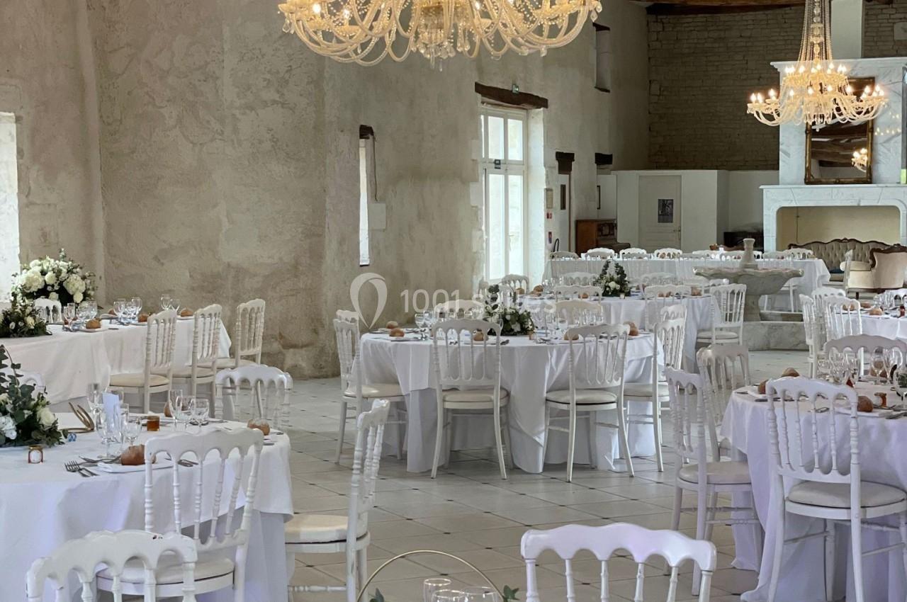 Salle de réception lumineuse avec tables décorées de fleurs blanches, chaises blanches et lustres élégants.