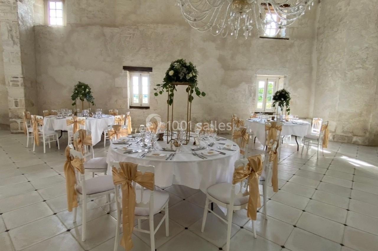 Salle de réception lumineuse avec tables rondes décorées de nappes blanches, chaises ornées de rubans dorés et centre de…