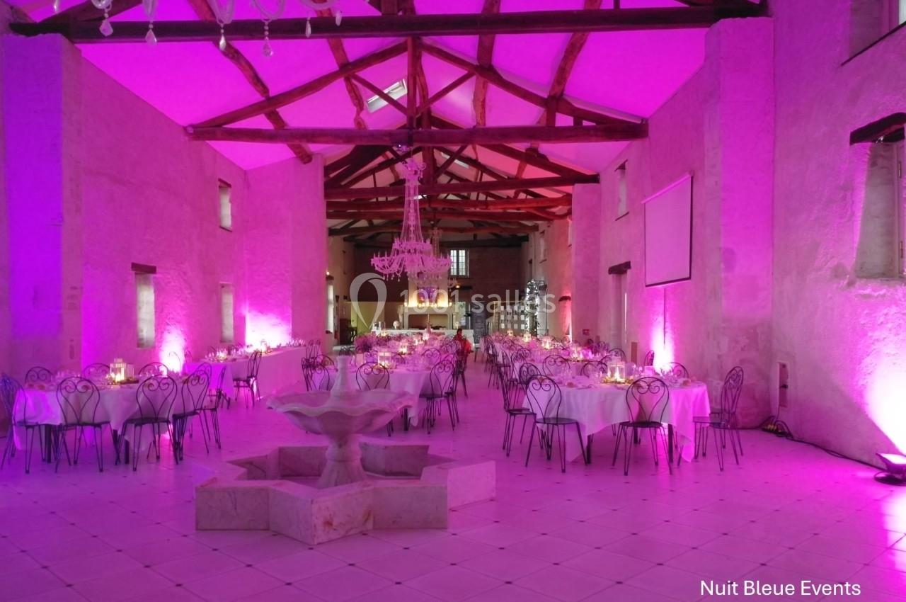 Salle de réception avec tables rondes dressées, éclairage rose, lustre central et fontaine en pierre au premier plan.